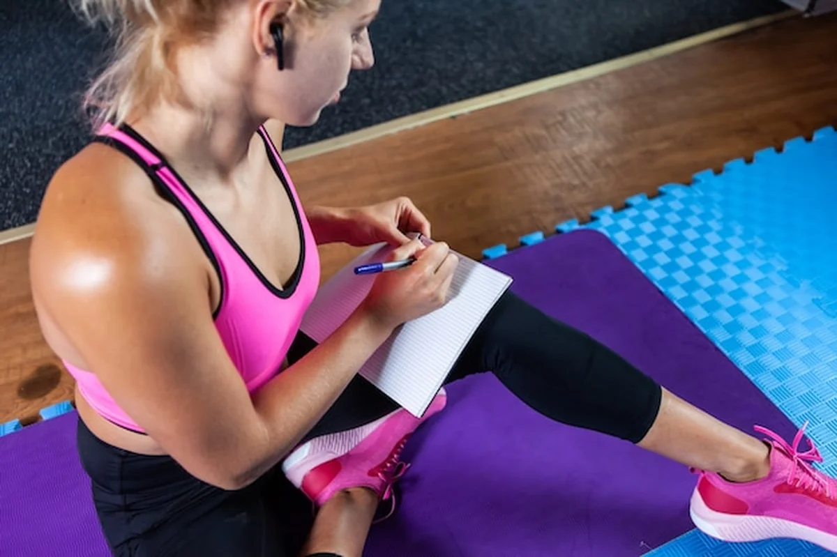 Blondynka fitness pisze w zeszycie treningowym na macie w klasie fitness